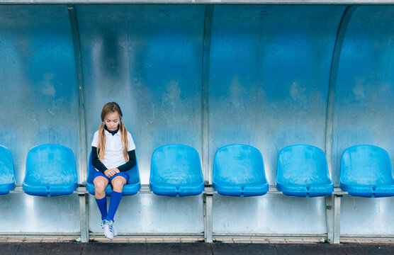 Full length frustrated preteen girl in soccer uniform sitting alone on blue plastic seat after match failure in sports club