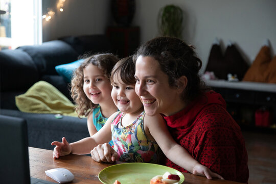 Cheerful young woman with cute little daughters sitting at table and enjoying video conversation with friends via laptop while spending evening at home