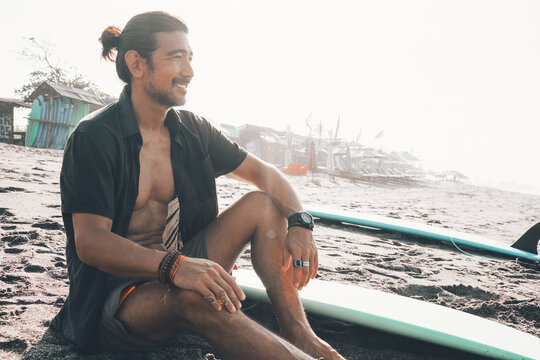 Positive young ethnic male in unbuttoned shirt and shorts enjoying summer day while resting on sandy beach near surfboard