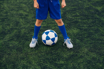 High angle of faceless child in blue shorts and cleats with ball standing alone on green court while playing football at modern stadium