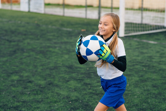 Cheerful Preteen Girl In White And Blue Uniform With Soccer Ball Smiling At Camera While Standing Alone On Green Field In Modern Sports Club