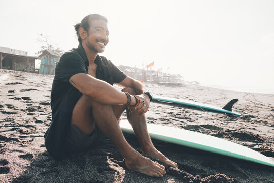 Positive young ethnic male in unbuttoned shirt and shorts enjoying summer day while resting on sandy beach near surfboard