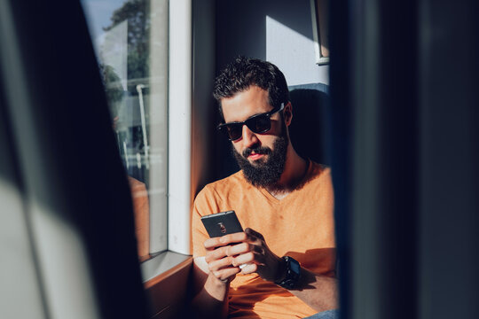 Calm bearded man in casual clothes and sunglasses using mobile phone while sitting on passenger seat near window in modern train
