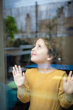Calm little girl with curly hair standing near window and looking away thoughtfully while spending time at home and dreaming about adventures
