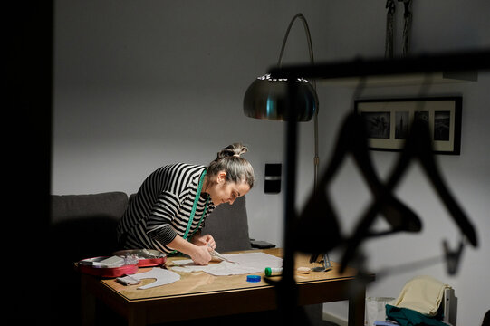 Side view of concentrated female designer in casual clothes standing near table and cutting fabric with scissors while working at home