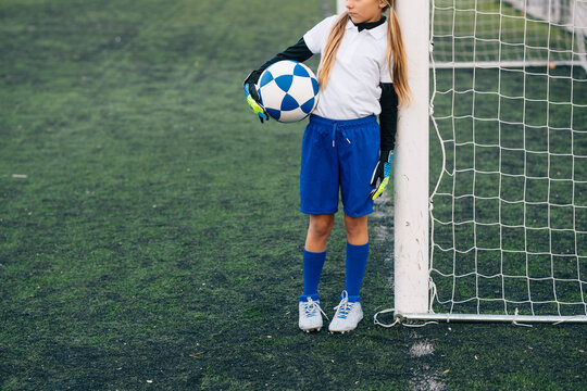 Crop girl in white and blue uniform with soccer ball while standing alone on green field in modern sports club