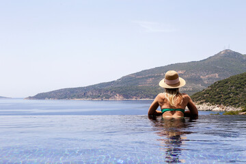 Young attractive woman enjoying the scenic ocean and mountain view from the edge of infinity pool on private villa. Back view of a sporty female in an outdoor swimming pool. Copy space, background.