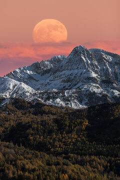 Majestic mountain landscape with full moon in colorful sky over snowy rocky range and dense forest in autumn time