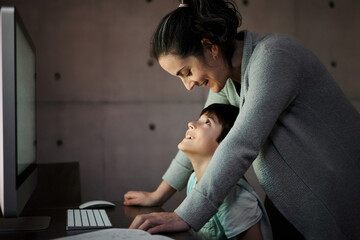 Side view of young woman explaining study task to positive son sitting at table with computer and textbook during online lesson at home
