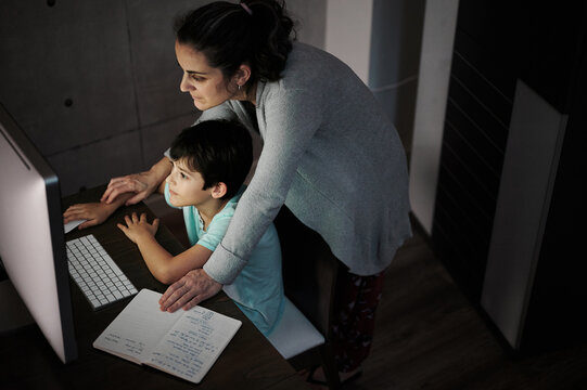 Side view of young woman explaining study task to positive son sitting at table with computer and textbook during online lesson at home