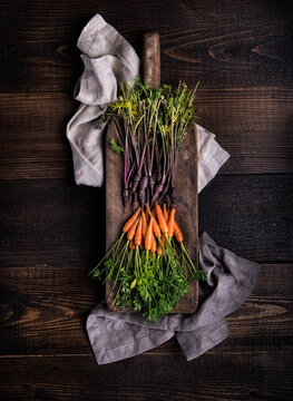 From above heaps of fresh purple and orange carrots on cutting board and natural fabrics on wooden rustic table
