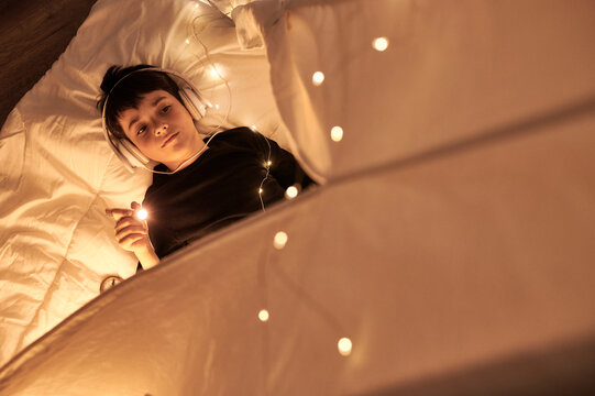 From above thoughtful male child in headset listening to music holding a flashlight lying in cozy kids playing tent in apartment