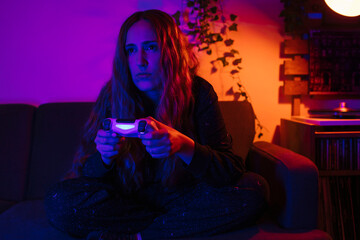 Young woman playing video games in her house during Corona Times. She is in a living room with colorful lights at night.