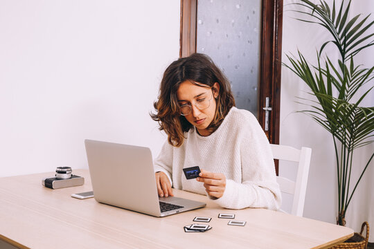 Young female photographer sitting at table with laptop and photo camera with projector and working with old photo slides at home workplace