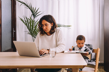 Young female freelancer in casual clothes sitting at table and working on project with laptop while little child sitting nearby with book