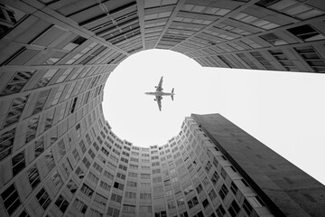 From below of black and white aircraft flying high over modern building with round yard in in city