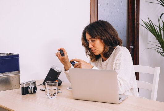 Young female photographer sitting at table with laptop and photo camera with projector and working with old photo slides at home workplace