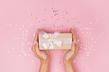Women hands holding a gift or gift box decorated with confetti on a pink pastel table top view