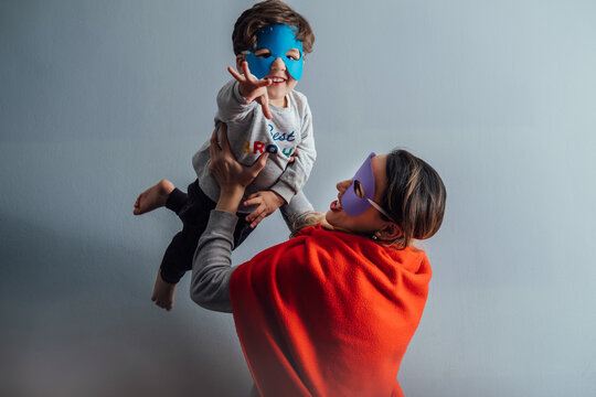 Side view of playful woman in superhero mask and cloak tossing little boy while spending time together at home and having fun