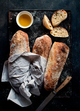 Ciabatta bread on rustic board near olive oil and knife with bread slices on dark background