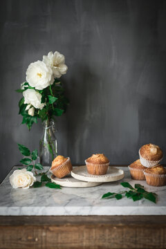 Still life of tasty yogurt cupcakes on round plates in arrangement with green leaves and aromatic white roses in glass vase on marble table against gray wall in kitchen