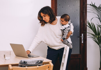Young busy female in casual wear with little kid on hand standing at table and checking email on laptop while working online at home