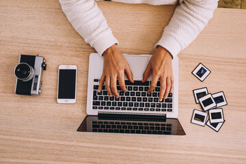 Top view of crop anonymous photographer typing on laptop keyboard while working with old photo slides at table with retro photo camera and modern smartphone