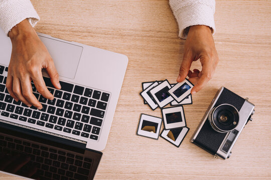 Top view of crop anonymous photographer typing on laptop keyboard while working with old photo slides at table with retro photo camera and modern smartphone