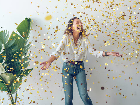 Beautiful young woman in stylish outfit laughing and looking up while standing under?falling golden confetti against white wall