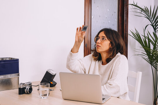 Young female photographer sitting at table with laptop and photo camera with projector and working with old photo slides at home workplace
