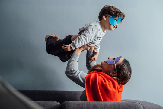 Side view of playful woman in superhero mask and cloak tossing little boy while spending time together at home and having fun