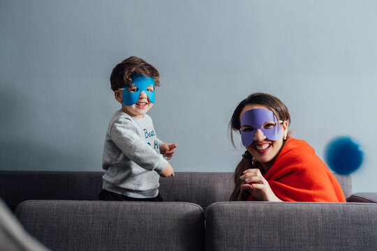 Cheerful woman and little boy wearing paper masks playing during weekend and pretending being superheroes while looking at camera