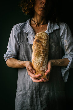 Cropped unrecognizable woman in apron holding ciabatta bread while standing on dark background