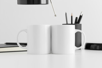 Two white mugs mockup with a lamp, notebook and workspace accessories on a white table.