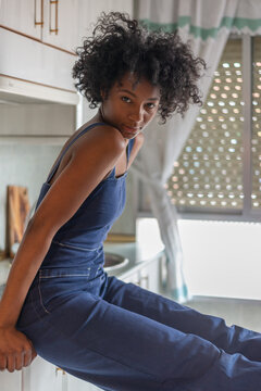 Side view of thoughtful exhausted black housewife with afro hairstyle sitting on counter and relaxing during housework while looking at camera near windows