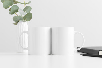 Two mugs mockup with eucalyptus in a vase and workspace accessories on a white table.