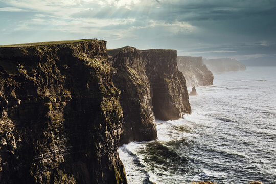 Perspective view of gigantic cliffs and waves dashing against remote wild coast of Ireland in sunbeams from cloudy sky