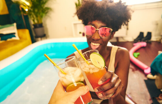 Happy black female in sunglasses and bikini sitting with alcohol drink garnished with slice of lime and relaxing near inflatable pool while staying at home