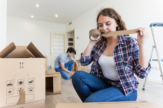 Young female cutting duct tape with teeth sitting near carton boxes and male sitting near black box in empty apartment