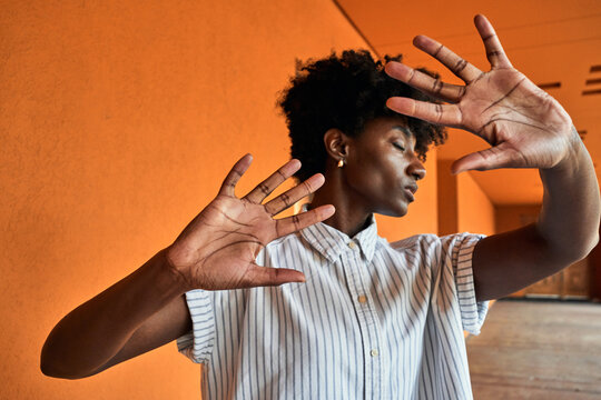 Adult African American female with eyes closed trying to stop negative impact while standing with arms raised against blurred vivid orange interior in corridor of modern building