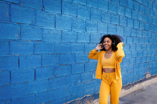 Satisfied African American female in bright clothes dancing with closed eyes near blue brick wall and enjoying songs in wireless headphones