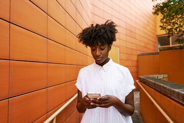 Adult African American female in white casual shirt messaging on smartphone standing on street against vibrant orange tiled facade of contemporary building in downtown