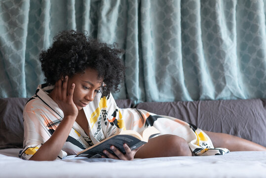 Content African American female in silk robe lying on comfortable bed and enjoying novel while leaning on hand