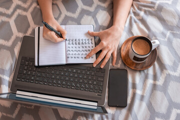 Top view of crop woman taking notes in bed while working next to his laptop, smartphone and cup of coffee