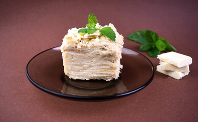 Piece of layered cake on a plate with mint leaves and pieces of white chocolate on background. Russian cake Napoleon on brown background. Birthday cake.