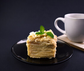 Piece of layered cake on a saucer with white cup on background. Russian cake Napoleon on dark background. Birthday cake. Space for text.