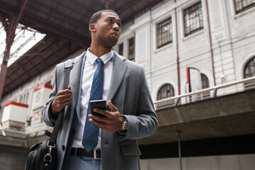 tHandsome black businessman in coat and with phone walking down moving stairs while commuting.
