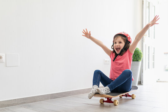 Little kid in red spotted helmet and casual clothes and dreaming about outdoors amusement while sitting alone on longboard in corridor of light modern apartment staying at home