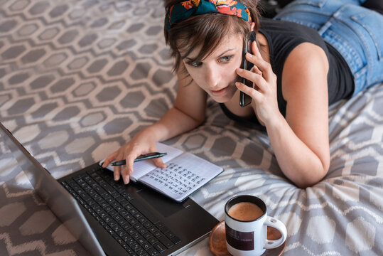 High angle of self employed female in casual wear lying on bed with laptop while speaking on mobile phone and taking notes in notebook