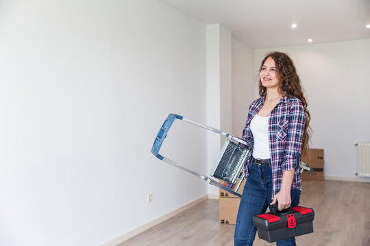 Young cute female standing in empty room with carton boxes with a ladder and a toolbox
